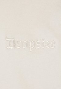 Tissu blanc cassé avec un logo "Dropsize" brodé dans une police avec empattement, pr�ésentant des points de couture en relief et texturés sur un fond lisse.