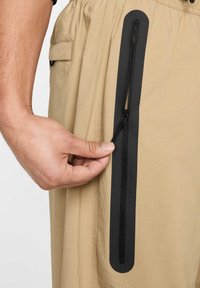 Shorts beige avec une poche zippée sur le côté dotée d'un zip noir et d'une languette. Le matériau semble léger avec une texture lisse.