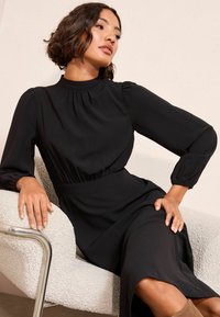 Melna garām piedurknēm kleita ar augstu kakla izgriezumu un savilkuma detaļu plecos, ar pieguļošu jostasvietu un plūstošu svārku.