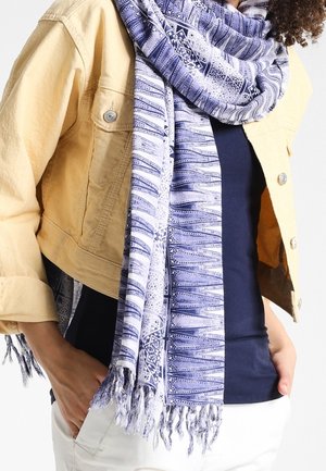 Femme portant une veste jaune, une chemise marine, un pantalon blanc et une écharpe bleue et blanche à motifs avec franges autour du cou.