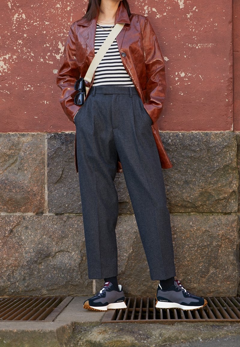 Personne portant un manteau en cuir marron, une chemise rayée, un pantalon gris taille haute, des baskets noires, et portant un sac bandoulière beige avec des lunettes de soleil.