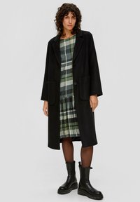 Schwarzer Wollmantel mit Reverskragen und zwei vorderen Taschen, getragen über einem grün karierten Kleid. Das Modell trägt schwarze Knöchelstiefel und transparente Strumpfhosen.