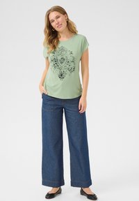 T-shirt vert clair à manches courtes avec un design graphique noir orné, associé à un jean décontracté en denim foncé à jambes larges et des chaussures plates noires.