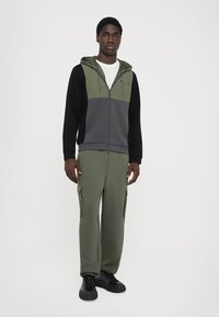Sweatshirt com fecho e puxadores ajustáveis em verde-oliva e cinza, com mangas pretas e bolsos laterais. Usado com calças cargo combinando.