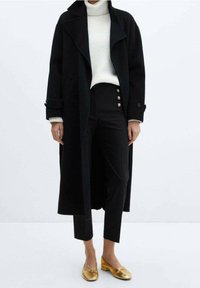 Manteau en laine noir à larges revers, associé à un col roulé blanc à côtes et à un pantalon noir taille haute avec des boutons dorés. Chaussures plates dorées.