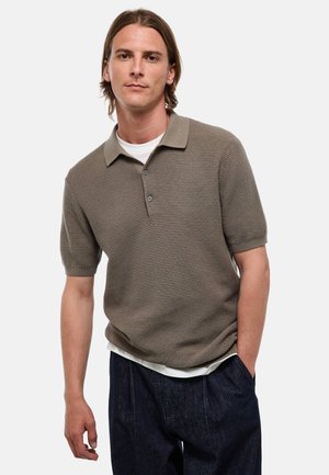 Homme aux cheveux lisses, mi-longs, porte un polo taupe texturé sur un t-shirt blanc et un pantalon foncé, debout avec une main dans la poche.
