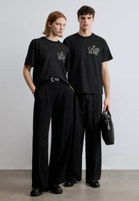 Μαύρα βαμβακερά T-shirts με χρυσαφί λογότυπο, συνδυασμένα με μαύρο παντελόνι φαρδιάς γραμμής. Η ανδρική φιγούρα κρατά μια μαύρη τσάντα, όλα σε ένα μινιμαλιστικό περιβάλλον.