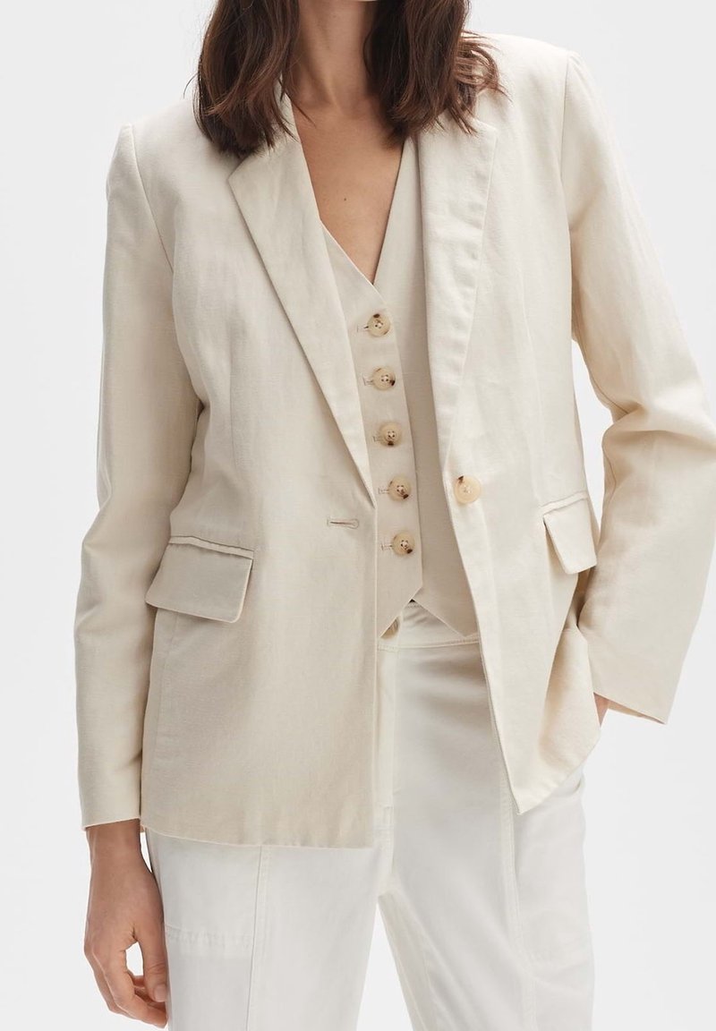 Femme portant un costume couleur crème avec blazer, gilet boutonné et pantalon assorti, une main dans la poche, sur un fond uni.