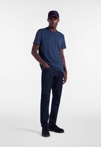 T-shirt navy con collo a giro, jeans in denim scuro e stivali in suede navy. La persona indossa un cappellino navy coordinato, in piedi su uno sfondo neutro.