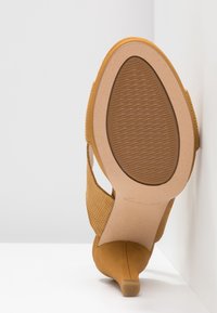 Semelle d’une chaussure à talons hauts couleur bronzée, inclinée verticalement contre un mur d’angle blanc montrant une zone de grip texturée et un talon fin.