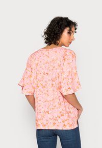Blusa floral em tecido rosa claro, com um corte solto, mangas curtas com folhos e pequenos padrões de flores em tons de laranja e amarelo.