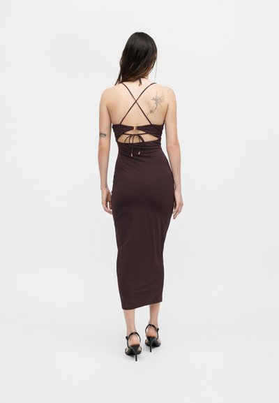 Γυναίκα που φοράει αμάνικο, ανοιχτή πλάτη, σκούρο καφέ midi φόρεμα με χιαστί τιράντες και τακούνια, στέκεται γυρισμένη προς τα πίσω σε μονόχρωμο φόντο.