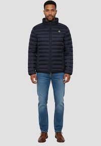 Veste matelassée bleu marine avec un col montant, un motif matelassé horizontal, et des poches zippées, portée avec un jean bleu clair et des chaussures marron.