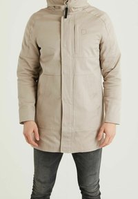 Man som bär en beige huvad jacka med dragkedja och fickor, tillsammans med mörkgrå åtsittande jeans, stående mot en helt enkel bakgrund.