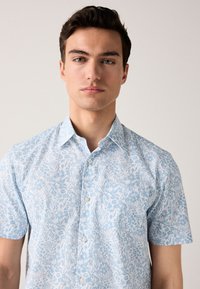 Világoskék, rövid ujjú, gombos ing fehér virágmintával, széthajtott gallérral és sima fehér gombokkal, könnyű anyagból készült.