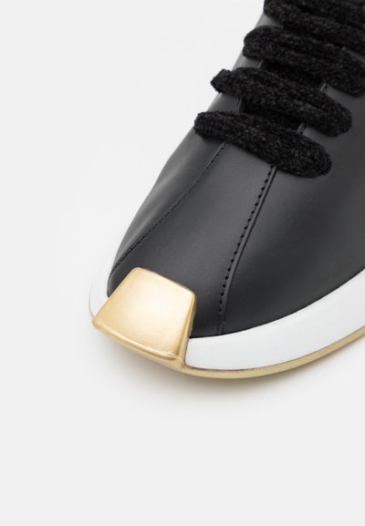 Sapatilhas de couro preto com uma biqueira dourada proeminente, atacadores texturizados pretos e uma entressola branca. Apresentam um design elegante e moderno.