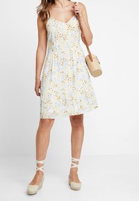 Blommig sommarklänning med v-ringning, gula och blå blommönster, figursydd överdel, utställd kjol och justerbara spagettiband. Vävd axelremsväska.