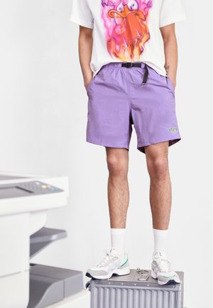 Persoon met paarse korte broek, witte sneakers en een T-shirt met opdruk die staat op een zilveren koffer naast een kantoorprinter.