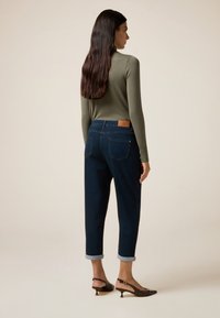 Jeans de mezclilla oscura con puños doblados, que cuentan con un parche de cuero marrón en la parte trasera. Llevados con una camiseta ajustada de manga larga verde y zapatos marrones.