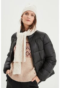 Giacca a vento nera sopra un maglione lavorato a maglia beige con motivi geometrici, sciarpa crema e berretto a maglia coordinato. Materiali lisci e strutturati.