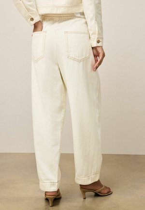 Persoană purtând pantaloni largi de culoare crem deschis, cu buzunare în spate, și sandale bej cu toc, stând pe un podea maro deschis.