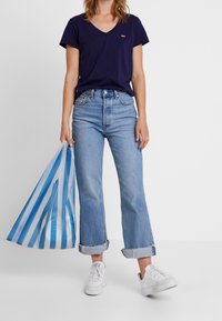 Marinblå V-ringad t-shirt med en liten logotyp, ljusblå vida jeans, uppvikta avslut och vita sneakers; håller en blå och vit randig väska.