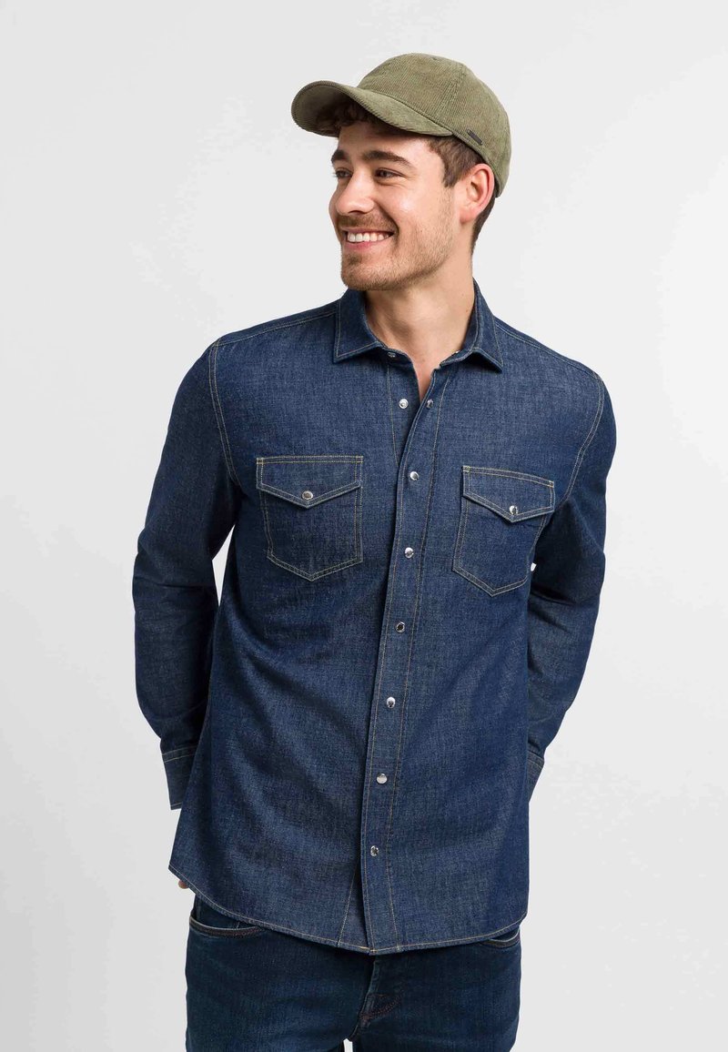 Dark denim shirt with buttoned front, two chest pockets, and contrasting stitching. Paired with a green cap, displayed on a neutral background.