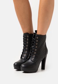 Botines negros de tacón alto con detalles texturizados, que presentan ojales metálicos, cordones delanteros y un acabado liso similar al cuero.