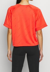 Jasnopomarańczowa sportowa koszulka z krótkim rękawem wykonana z gładkiej tkaniny, z subtelnym wzorem z logo i luźnym krojem.