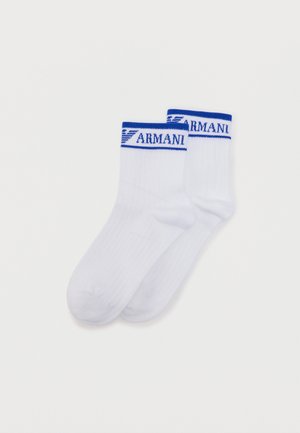 Pár bielych ponožiek s modrým lemom, na ktorom je logo Armani a emblém orla, na jednoduchom pozadí.