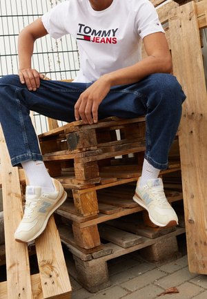 Osoba siedząca na ułożonych drewnianych paletach, ubrana w białą koszulkę Tommy Jeans, niebieskie dżinsy, białe skarpetki i beżowe sneakersy New Balance.