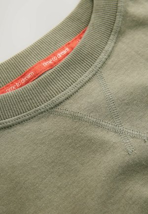 Olivgrön sweatshirtkrage med ribbad textur; flatlock-sömmar och en röd innermärke som visar "dags att drömma."