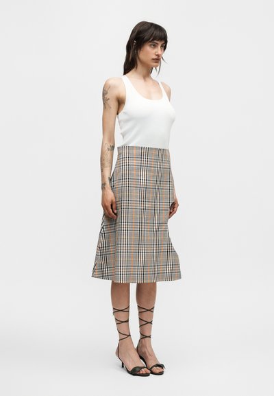 Γυναίκα με σκουρόχρωμα μαλλιά μέχρι τον ώμο φοράει λευκό μπλουζάκι χωρίς μανίκια, καρό φούστα μέχρι το γόνατο και μαύρα δερμάτινα παπούτσια με κορδόνια, στέκεται μπροστά σε μονόχρωμο φόντο.