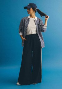 Pasiasta koszula z guzikami w niebiesko-białe pasy, zestawiona z czarnymi spodniami o szerokim kroju z białymi paskami po bokach. Całość dopełnia granatowa czapka bejsbolowa.