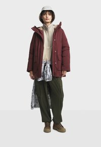 Bordeaux kleurige pufferjas met twee zakken, olijfgroene cargobroek, beige fleece top en een grijze bucket hat. Patroon op het geknoopte shirt is zichtbaar. Bruine laarzen.