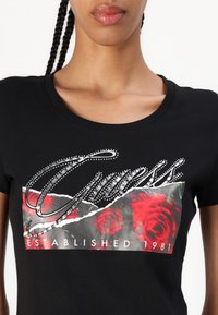 Black t-shirt featuring a graphic print with red roses and a silver "Guess" logo embellished with rhinestones. Crew neck design.