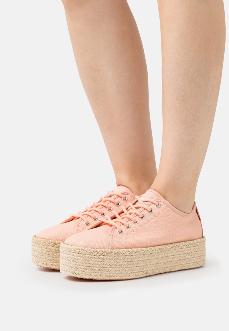 Jambes portant des baskets compensées en toile rose avec des semelles en jute beige épaisses, se tenant sur une surface blanche neutre.