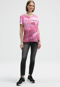Young woman standing wearing pink "HAPPY TIME" t-shirt, faded black jeans with text, and black sneakers against plain white background.