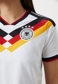 Vrouw die een wit Duits voetbalshirt draagt met zwarte, rode en gele chevronstrepen en het embleem van de Deutscher Fussball-Bund op de borst.