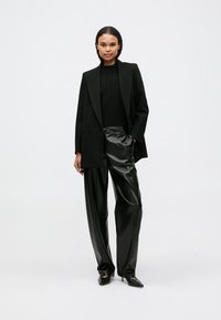 Blazer preto ajustado sobre um top preto canelado, combinado com calças pretas de imitação de couro brilhantes de cintura alta e sapatos de salto pretos pontiagudos.