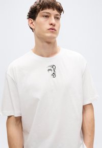 Λευκό βαμβακερό T-shirt με κοντά μανίκια, που διαθέτει μικρό μαύρο φυλετικό σχέδιο στο στήθος. Λεία υφή και άνετη εφαρμογή.