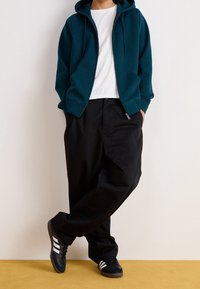 Teal zip-up hoodie, vit t-shirt, svarta lösa byxor och svarta sneakers med vita ränder; stående på en gul yta.