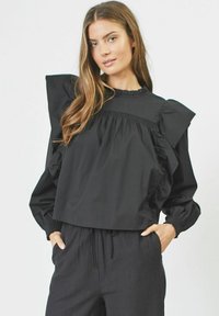 Blusa cropped nera con maniche a volant, polsini elasticizzati e scollatura arricciata. Indossata con pantaloni neri larghi. Tessuto dalla consistenza liscia.