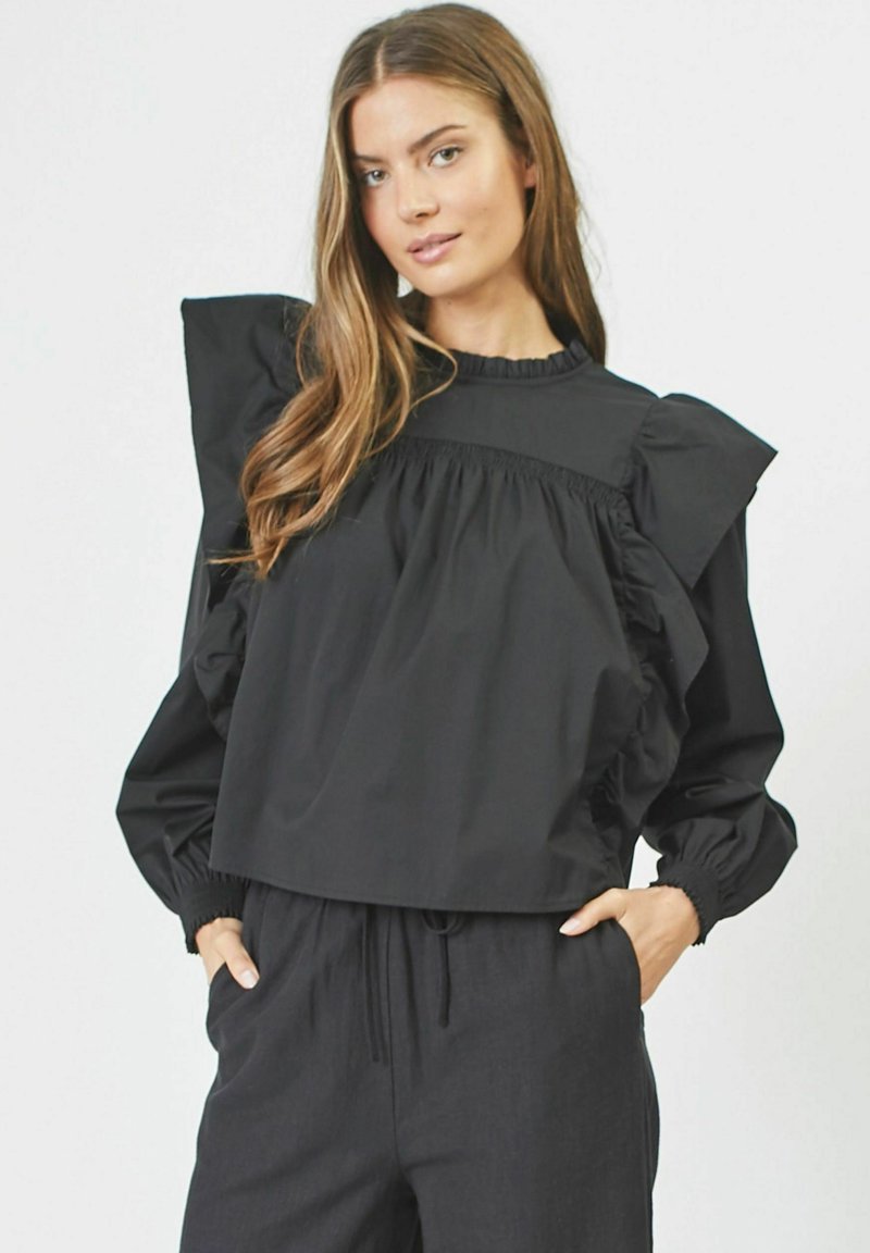 Blusa cropped nera con maniche a volant, polsini elasticizzati e scollatura arricciata. Indossata con pantaloni neri larghi. Tessuto dalla consistenza liscia.
