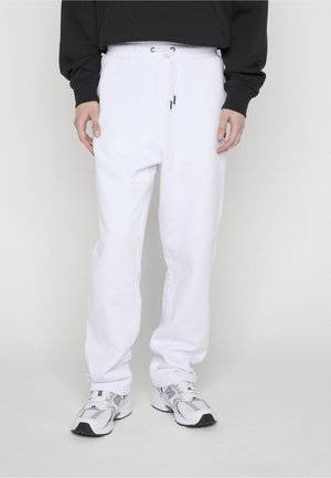 Person trägt weiße Sweatpants mit Kordelzug, schwarzen Langarmshirt und weiße New Balance Sneakers und steht vor einem schlichten Hintergrund.