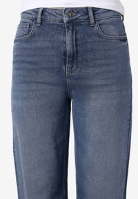 Γυναικείες τζιν παντελόνες από μπλε τζιν, με μπροστινές τσέπες, θηλιές ζώνης, κουμπί και φερμουάρ, φορεμένες με άσπρη μπλούζα.