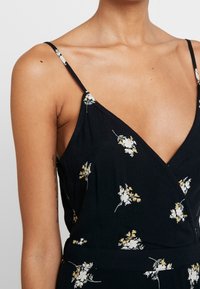 Mono floral negro con tirantes finos, que presenta patrones de flores blancas y amarillas, un diseño de envoltura y una tela texturizada.