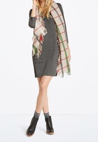 Robe tricotée grise à manches longues, avec poches avant ; ornée d'une écharpe à franges en plaid crème, vert et rouge. Bottes noires à la cheville et chaussettes grises.