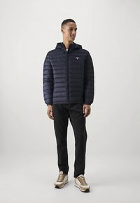 Marineblå dynejakke med vandret syning, lynlås foran og hætte. Kombineret med sorte jeans og hvide sneakers med gyldne accenter.