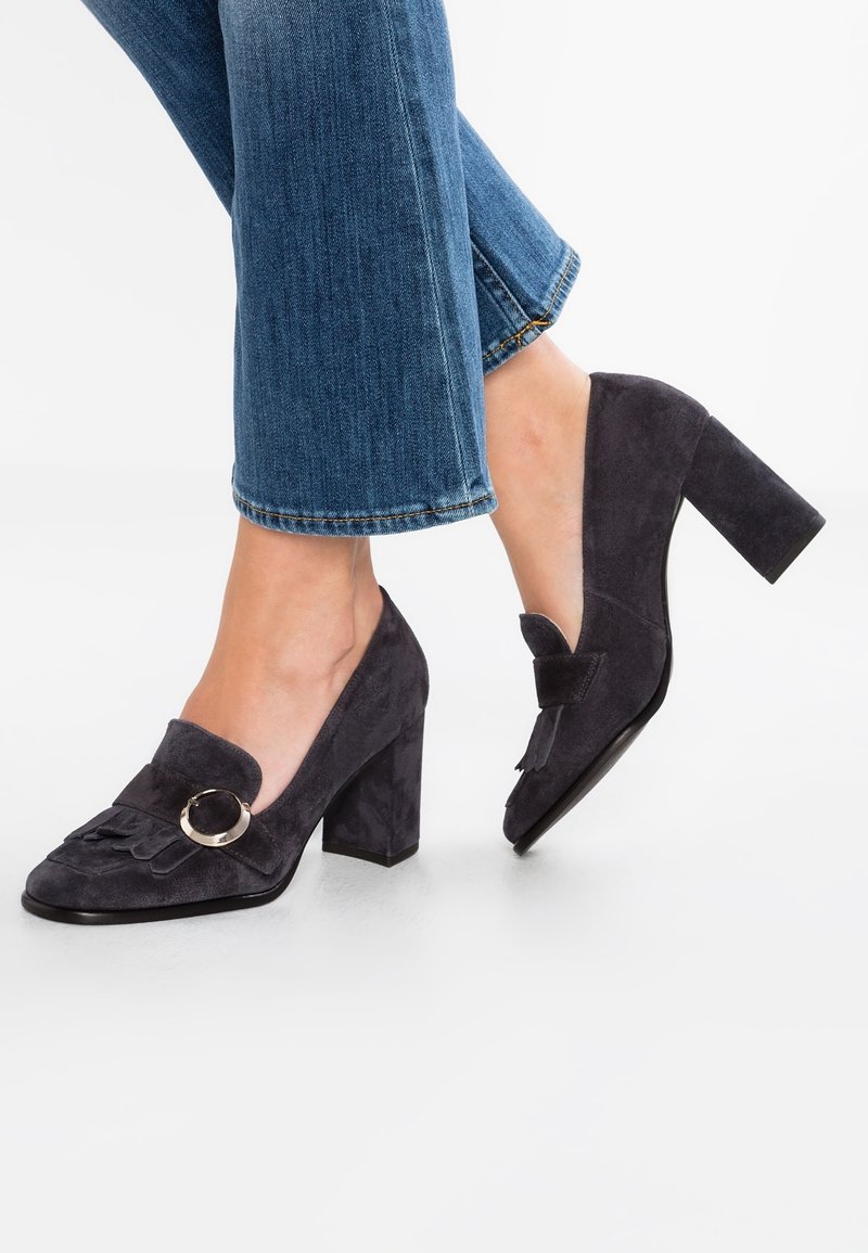 Chaussures à talon bloc en daim gris foncé avec un bout rond, des franges décoratives et un accent métallique circulaire à l'avant.